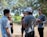 A group of young people is engaged in a conversation in a park-like setting. They stand under the shade of tall trees with sunlight filtering through the leaves. One person is holding a football, suggesting a casual or recreational gathering.