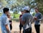 A group of young people is engaged in a conversation in a park-like setting. They stand under the shade of tall trees with sunlight filtering through the leaves. One person is holding a football, suggesting a casual or recreational gathering.