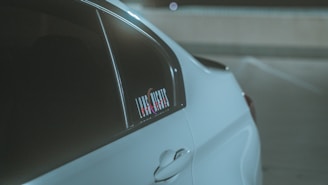 A close-up view of a sleek white car's rear side panel, emphasizing its smooth curves and window. A sticker with text is visible on the window. The scene appears to be in a dimly lit parking area.