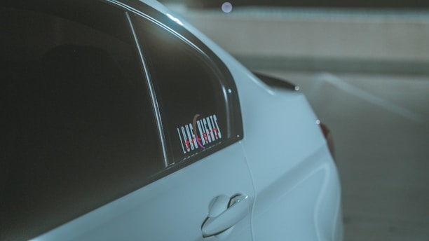 A close-up view of a sleek white car's rear side panel, emphasizing its smooth curves and window. A sticker with text is visible on the window. The scene appears to be in a dimly lit parking area.