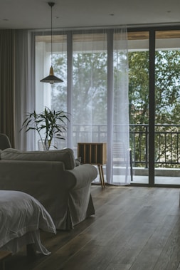 A cozy living room with a neutral-colored sofa and a potted plant in front of large glass windows. Sheer white curtains partially cover the windows, allowing natural light to fill the space. A modern pendant light hangs from the ceiling, and there is a small wooden side table next to the sofa. A balcony area with chairs and greenery outside complements the interior.