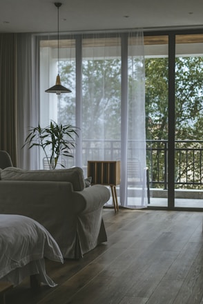 A cozy living room with a neutral-colored sofa and a potted plant in front of large glass windows. Sheer white curtains partially cover the windows, allowing natural light to fill the space. A modern pendant light hangs from the ceiling, and there is a small wooden side table next to the sofa. A balcony area with chairs and greenery outside complements the interior.