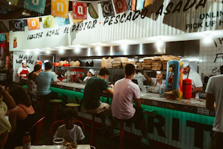 A friendly staff member taking an order at Los Takitos counter with colorful taco posters in the background.
