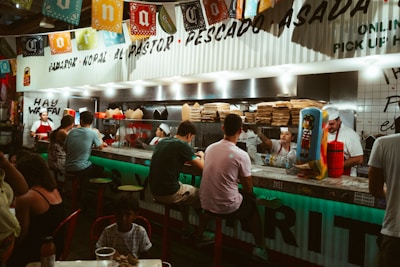 A lively food stall or taqueria with a row of people seated on stools at a counter. There are colorful paper banners hanging from the ceiling and several staff members behind the counter preparing food. The stall has a vibrant atmosphere with various signs and decorations, including stacks of brown bags and a drink dispenser. The scene shows a group of customers enjoying their meals and drinks.