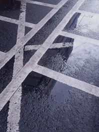 Pavement with fresh rain reflecting the sky, showing white painted lines forming boxes and intersections. Water droplets create a textured, reflective surface on the dark asphalt.