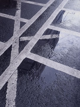 Pavement with fresh rain reflecting the sky, showing white painted lines forming boxes and intersections. Water droplets create a textured, reflective surface on the dark asphalt.