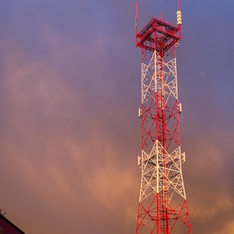 A tall red and white radio or telecommunication tower stands against a backdrop of a dramatic sky. The structure is composed of interconnecting steel beams with antennas attached at various levels.