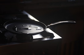 A frying pan with small puddles of oil sits on a gas stove, illuminated by soft, directional light that highlights its glossy surface. The background is dark, creating a dramatic contrast with the highlighted pan.