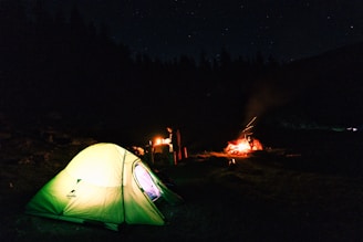Campfire glowing at dusk with a handmade shelter in the background.