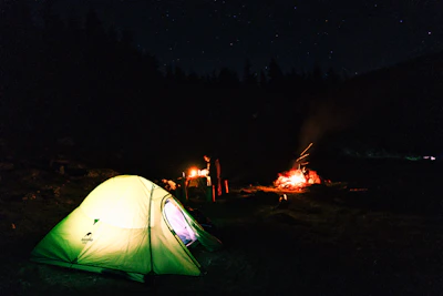 A cozy campsite setup with a glowing tent, campfire, and lush forest background at dusk.