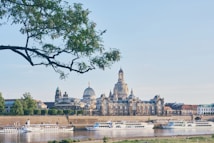 A scenic view featuring an ornate baroque building with a prominent dome and spires, set along a riverbank. Several river cruise ships are docked in the foreground. A large leafy tree branch frames the upper part of the image, and the sky is clear and blue.