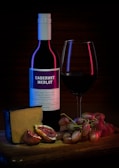 A bottle of Cabernet Merlot wine with a filled glass sits on a wooden surface. Nearby are a wedge of cheese, halved figs, and a bunch of grapes. Soft lighting highlights textures and colors, giving a sense of luxury and indulgence.