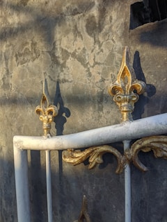 The image contains a decorative wrought iron fence or railing against a textured concrete wall. The metal features intricate fleur-de-lis patterns with a mix of gold and white colors. Shadows of nearby objects are cast onto the wall, creating a dramatic effect.