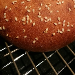 A close-up view of a freshly baked bun with a brown crust, sprinkled with sesame seeds. The bun is resting on a metal cooling rack with visible grid patterns.