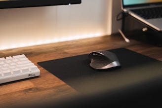 A modern workspace with a wooden desk featuring a white mechanical keyboard and an ergonomic mouse on a black mouse pad. A laptop is partially visible to the right, and the scene is softly lit with a warm ambient light.