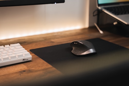A modern workspace with a wooden desk featuring a white mechanical keyboard and an ergonomic mouse on a black mouse pad. A laptop is partially visible to the right, and the scene is softly lit with a warm ambient light.