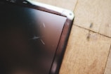 A close-up view of a scratched tablet or phone screen next to a wooden surface. The screen shows multiple scratches and the wooden surface has a natural grain pattern.