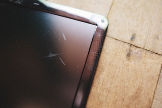 A close-up view of a scratched tablet or phone screen next to a wooden surface. The screen shows multiple scratches and the wooden surface has a natural grain pattern.