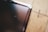 A close-up view of a scratched tablet or phone screen next to a wooden surface. The screen shows multiple scratches and the wooden surface has a natural grain pattern.
