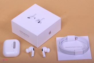 A set of white wireless earbuds is displayed alongside the packaging. The items include two individual earbuds, a compact charging case, a neatly coiled charging cable, and a white square packaging box labeled 'AirPods Pro'. The background is a neutral beige color.