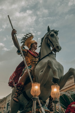 A statue of a warrior wearing golden armor with a feathered helmet, holding a spear, and mounted on a rearing horse. The scene includes ornate street lamps and a backdrop of a cloudy sky with some buildings visible.