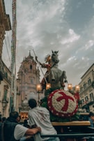 A festive procession is depicted with a large statue of a horse and rider, possibly a saint, adorned with intricate decorations and surrounded by people. The background features an ornate building or church under a cloudy sky.