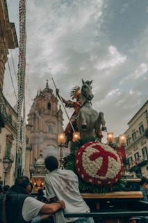 A festive procession is depicted with a large statue of a horse and rider, possibly a saint, adorned with intricate decorations and surrounded by people. The background features an ornate building or church under a cloudy sky.