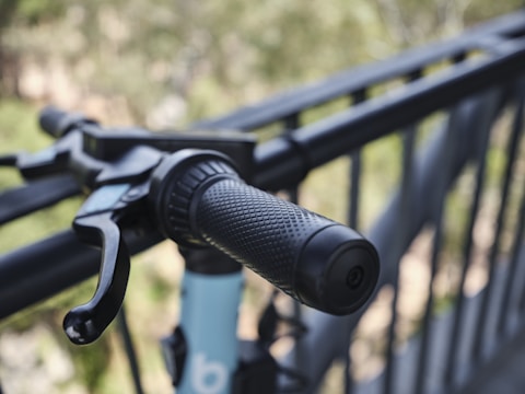 Close-up view of a bicycle handlebar with a textured rubber grip and a brake lever, set against a blurred background of a metal railing and greenery.