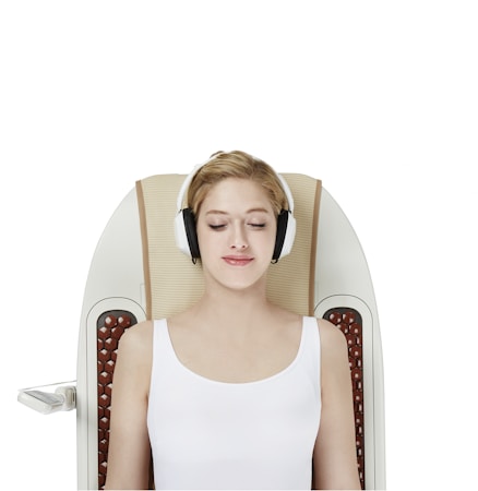 A woman with headphones is lying on a massage chair with her eyes closed. She is wearing a white sleeveless top and appears to be relaxed. The chair has a sleek, modern design with padded sections and control panels on the side.