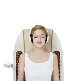 A woman with headphones is lying on a massage chair with her eyes closed. She is wearing a white sleeveless top and appears to be relaxed. The chair has a sleek, modern design with padded sections and control panels on the side.