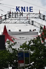 A large metal arch structure with the word 'KNIE' in bold blue letters is visible against a cloudy sky. Below, there are green leafy trees partially obscuring a circus tent with red peaks. A public transportation sign labeled 'Bellevue' appears in the foreground.