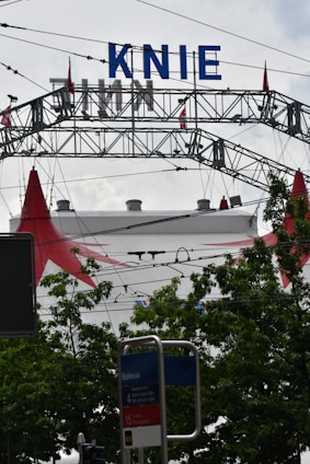 A large metal arch structure with the word 'KNIE' in bold blue letters is visible against a cloudy sky. Below, there are green leafy trees partially obscuring a circus tent with red peaks. A public transportation sign labeled 'Bellevue' appears in the foreground.
