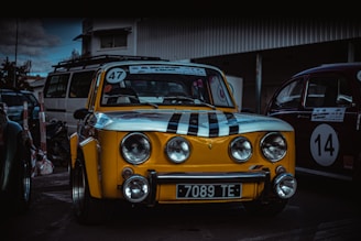A vintage yellow car with black racing stripes and the number 47 displayed on the windshield. The car is parked in an outdoor area alongside other classic vehicles, such as a red car with the number 14. The setting appears to be a parking lot or a garage area with overcast skies.