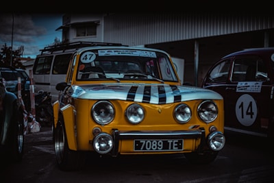 A vintage yellow car with black racing stripes and the number 47 displayed on the windshield. The car is parked in an outdoor area alongside other classic vehicles, such as a red car with the number 14. The setting appears to be a parking lot or a garage area with overcast skies.