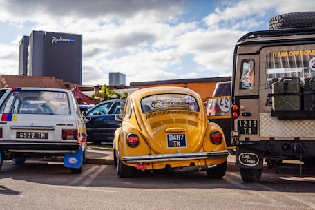 Three vintage cars, including a yellow Volkswagen Beetle, are parked closely together in a lot. A tall building in the background with the 'Radisson Blu' logo indicates an urban setting. The sky is partly cloudy.
