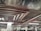 A parking garage features rows of parked cars beneath a complex network of metallic ventilation ducts on the ceiling. The ceiling also has a reflective surface, and the area is lit with fluorescent lights.