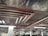 A parking garage features rows of parked cars beneath a complex network of metallic ventilation ducts on the ceiling. The ceiling also has a reflective surface, and the area is lit with fluorescent lights.