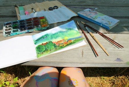 A watercolor painting in a sketchbook depicting a vibrant landscape scene with trees, a house, and mountains. The setup includes a wooden table with watercolor paints, brushes, and drawing supplies. Sunlight casts gentle shadows across the table, and the artist's legs are visible, decorated with colorful patterns.