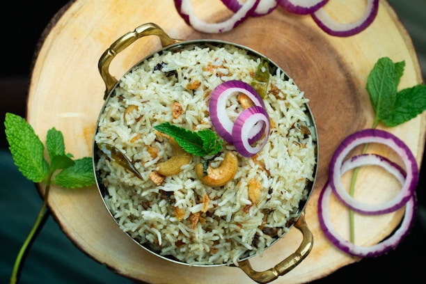 A dish of rice garnished with sliced onions, mint leaves, and cashews, served in a brass bowl on a wooden surface. The presentation includes a decorative arrangement of red onion rings and fresh mint sprigs placed around the dish.