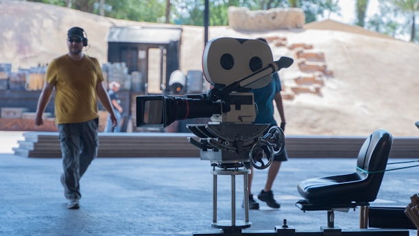 A film set with a vintage movie camera on a tripod in the foreground and two men walking in the background. The scene appears to be outdoors with a sandy backdrop and some wooden structures. There are also some chairs and various pieces of filming equipment visible.