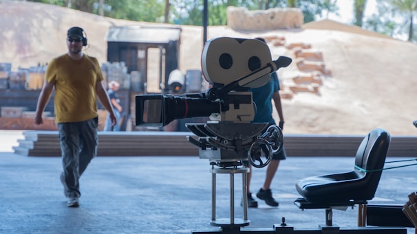 A film set with a vintage movie camera on a tripod in the foreground and two men walking in the background. The scene appears to be outdoors with a sandy backdrop and some wooden structures. There are also some chairs and various pieces of filming equipment visible.