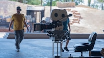 A film set with a vintage movie camera on a tripod in the foreground and two men walking in the background. The scene appears to be outdoors with a sandy backdrop and some wooden structures. There are also some chairs and various pieces of filming equipment visible.
