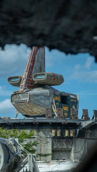 A futuristic spaceship with a red and gray body is docked on a platform surrounded by industrial structures. The ship features distinctive wing designs with large engine pods. In the background, a bright blue sky with fluffy clouds can be seen, contrasting with the metal and concrete elements of the foreground.