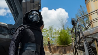 A person dressed in a futuristic black outfit with a helmet stands beneath a large metallic structure. The background features a bright blue sky with scattered clouds and some green foliage. There is a tall column on the right and a railing with several cables.