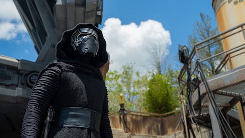 A person dressed in a futuristic black outfit with a helmet stands beneath a large metallic structure. The background features a bright blue sky with scattered clouds and some green foliage. There is a tall column on the right and a railing with several cables.