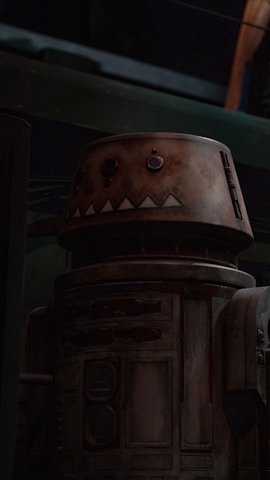 A vintage, weathered robot with a cylindrical body and a round head displaying rudimentary facial features is positioned within a dimly lit industrial setting. The surface of the robot is characterized by a rusty and rugged texture, suggesting age and wear.