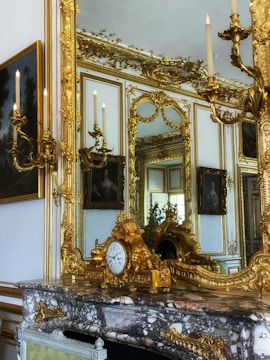 An opulent interior featuring an ornate mirror with elaborate golden frames and decorative sconces. A marble fireplace mantel holds a gilded clock adorned with cherubic figures. The room's walls are adorned with luxurious gold decorations, and there are classic paintings visible, reflecting a grand and luxurious setting.