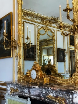 An opulent interior featuring an ornate mirror with elaborate golden frames and decorative sconces. A marble fireplace mantel holds a gilded clock adorned with cherubic figures. The room's walls are adorned with luxurious gold decorations, and there are classic paintings visible, reflecting a grand and luxurious setting.