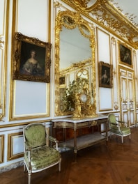 An opulent interior setting featuring ornate gold-trimmed walls, elaborate framed portraits, and a large decorative mirror. Between the portraits is an intricately carved wooden console topped with gilded decorations, and a pair of elegant green upholstered chairs flanking the console. The parquet wooden floor adds to the luxurious atmosphere.