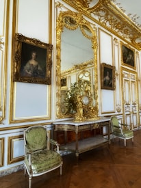 An opulent interior setting featuring ornate gold-trimmed walls, elaborate framed portraits, and a large decorative mirror. Between the portraits is an intricately carved wooden console topped with gilded decorations, and a pair of elegant green upholstered chairs flanking the console. The parquet wooden floor adds to the luxurious atmosphere.